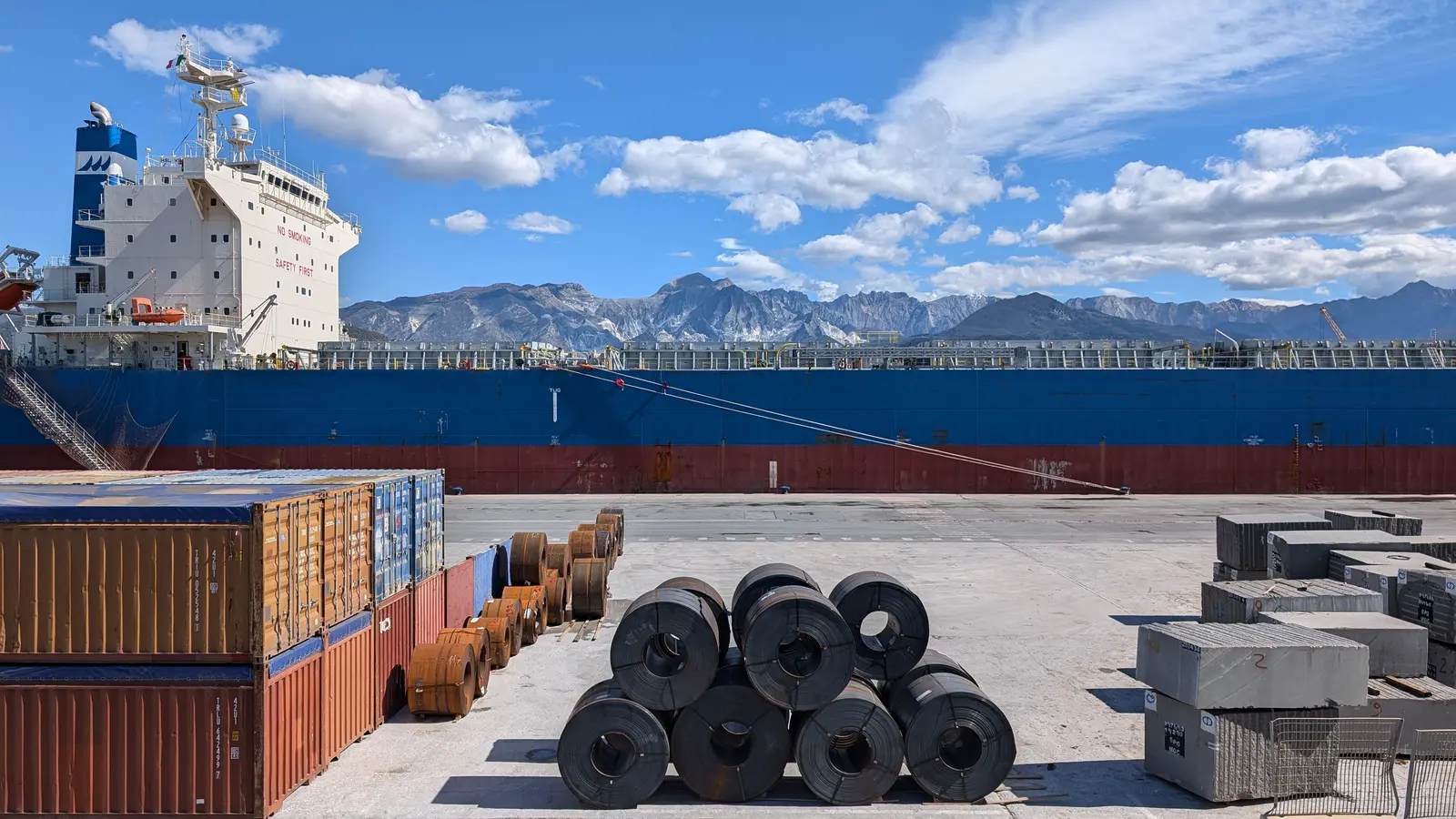 Warmgewalste staalrollen en containers op de kade naast een bulkschip in de haven van Carrara, Italië.